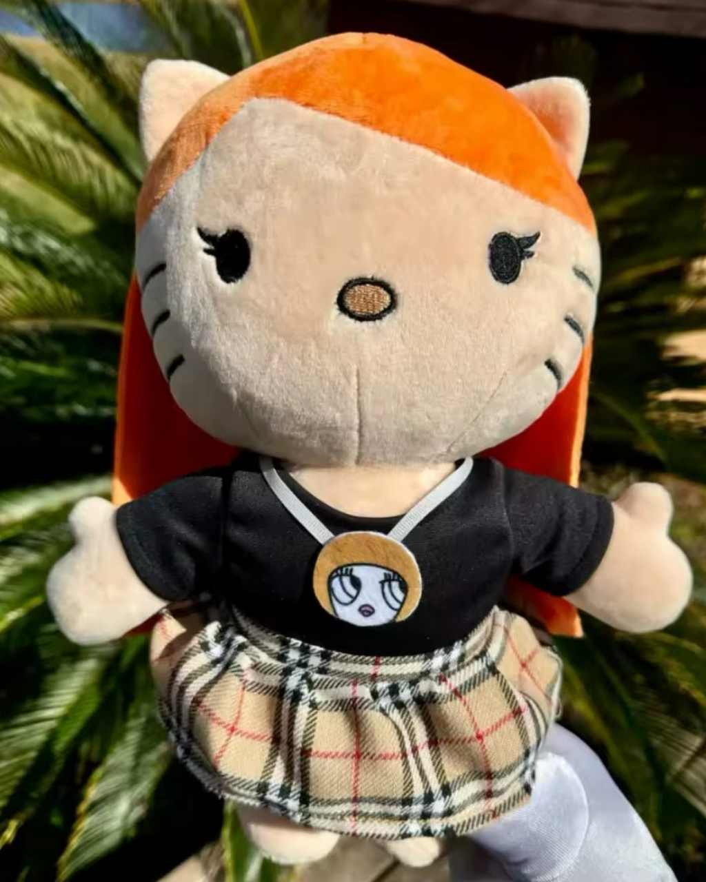 Ice Spice Hello Kitty plush in a plaid skirt photographed outdoors against green foliage.