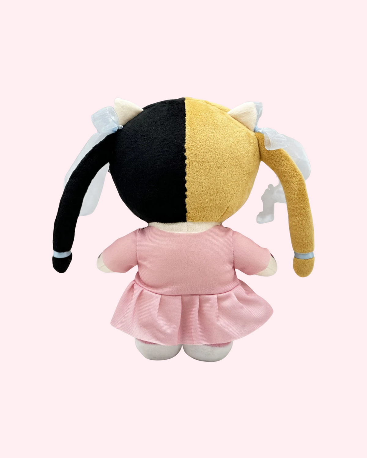 Back view of Melanie Martinez Hello Kitty plush with blond-and-black pigtails, a pink dress and white shoes, on a pale pink background.