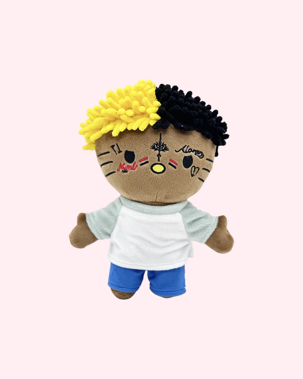 Front view of XXXTentacion Hello Kitty plush with yellow-and-black hair, embroidered face tattoos, a white shirt and blue shorts, on a pale pink background.